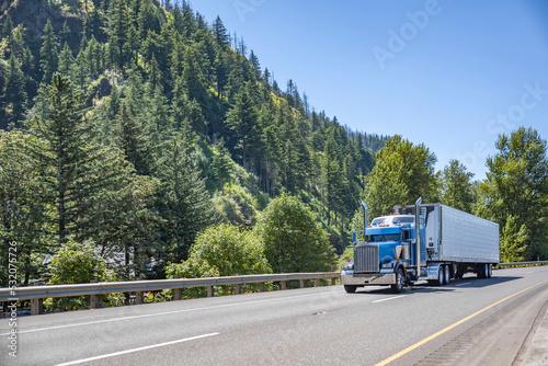 Blue American idol classic bonnet big rig semi truck tractor transporting frozen food in reefer semi trailer driving on the highway road with forest on the side