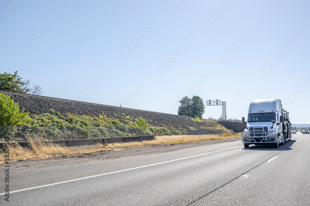 Industrial white big rig car hauler semi truck with high cab roof transporting cars on the semi trailer driving on the turning highway Industrial white big rig car hauler semi truck with high cab roof transporting cars on the semi trailer driving on the turning highway
