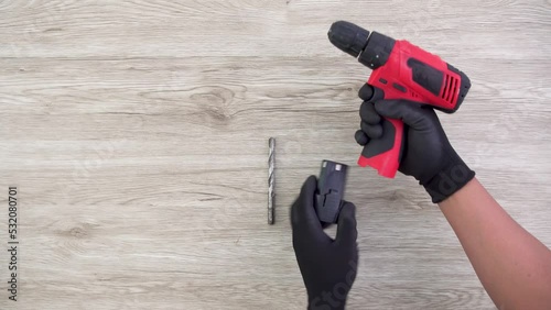 Two hand wearing black gloves attaching battery and drill bit to red drill with wooden background photo