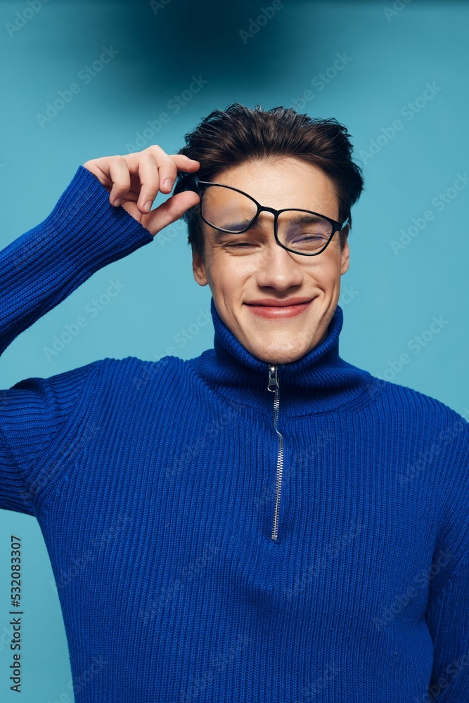 a handsome, attractive man stands on a light blue background in a blue zip-up sweater with black-rimmed glasses turned upside down on his face, looking at the camera with a charming look