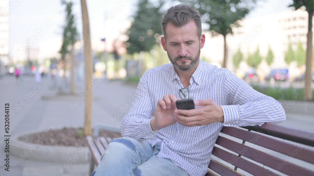 Young Adult Man Using Smartphone while Relaxing on Bench Young Adult Man Using Smartphone while Relaxing on Bench