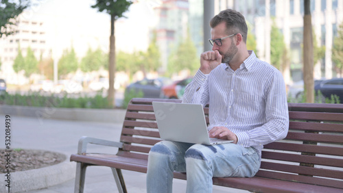 Coughing Young Adult Man Using Laptop while Sitting Outdoor on Bench © stockbakers