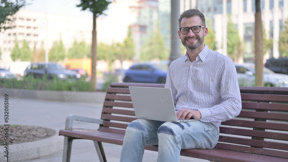 Young Adult Man with Laptop Smiling at Camera while Sitting Outdoor on Bench