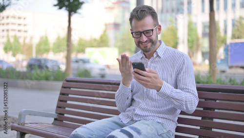 Young Adult Man Celebrating Online Success on Smartphone while Sitting Outdoor on Bench