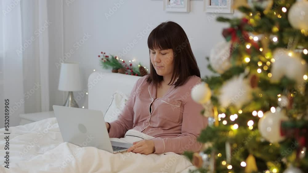 christmas concept - young woman in pajamas sitting on bed and using laptop near decorated christmas tree christmas concept - young woman in pajamas sitting on bed and using laptop near decorated christmas tree