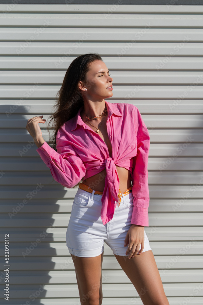 Young beautiful brunette woman in a pink shirt and white denim shorts  against the background of a light garage door fence Stock Photo | Adobe  Stock