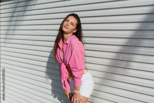 Young beautiful brunette woman in a pink shirt and white denim shorts against the background of a light garage door fence