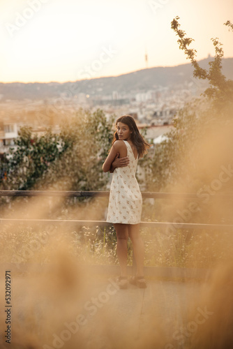 Beautiful young caucasian girl in white dress posing looking at camera outdoors at sunset. Brunette hugs herself with her arms  spends evening alone. Leisure lifestyle concept