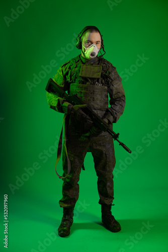 a soldier in the studio on a green background puts on a white mask on his face. man in military uniform with a rifle or machine gun