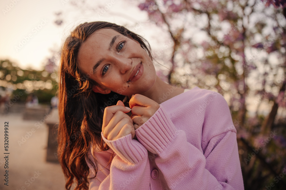 Attractive young caucasian woman looking at camera with her head bowed against blurred park background. Brunette with wavy hair wears pink sweater. Cozy spring concept Attractive young caucasian woman looking at camera with her head bowed against blurred park background. Brunette with wavy hair wears pink sweater. Cozy spring concept