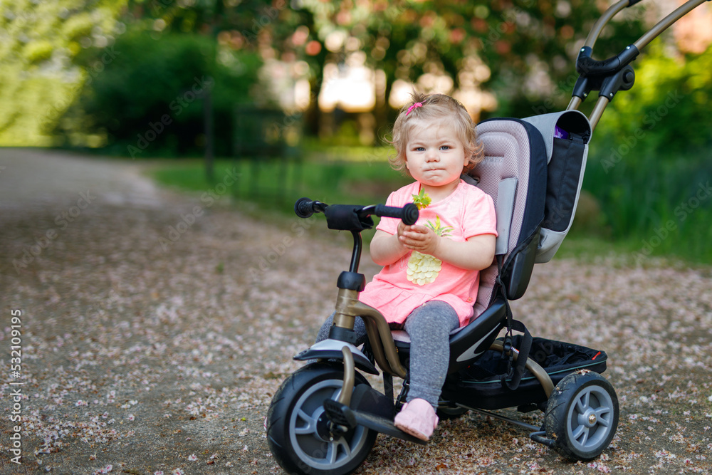 Cute adorable toddler girl sitting on pushing bicycle or tricycle. Little baby child going for a walk with parents on sunny day. Happy healthy kid in colorful clothes Cute adorable toddler girl sitting on pushing bicycle or tricycle. Little baby child going for a walk with parents on sunny day. Happy healthy kid in colorful clothes