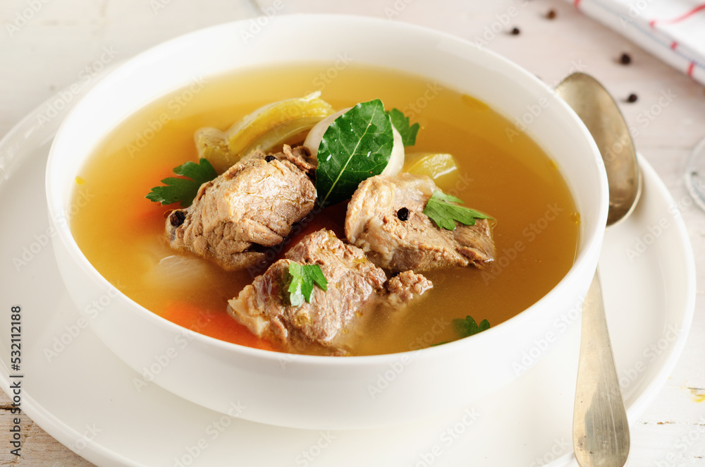 Autumn winter warming beef broth with a piece of meat and vegetables in a white dish on light background. Close up. Autumn winter warming beef broth with a piece of meat and vegetables in a white dish on light background. Close up.