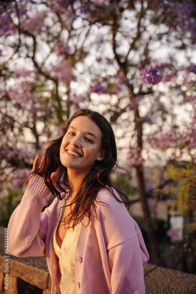 Outdoor portrait of young caucasian cute lady smiling posing near blossoming tree. Brunette with wavy hair wears pink sweater. Cozy spring concept Outdoor portrait of young caucasian cute lady smiling posing near blossoming tree. Brunette with wavy hair wears pink sweater. Cozy spring concept