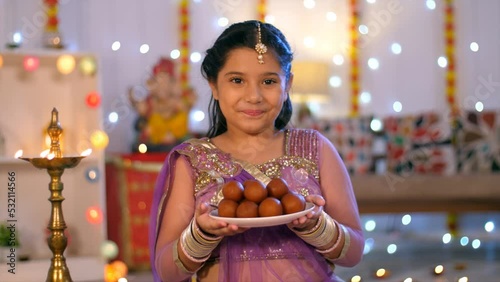 A sweet Indian girl offering Gulaab- Jamuns during Diwali celebration - traditional Indian sweets  Indian Diwali  Diwali decoration . Diwali lamp burning against a colorful decorated wall for Diwal... photo