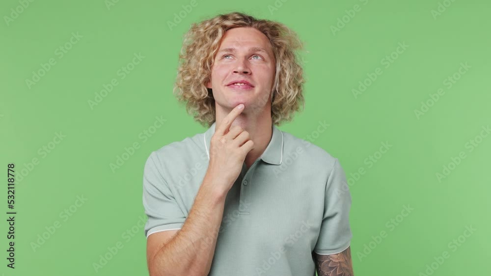 Young fun caucasian blond man 20s he wear blue t-shirt look around think dream put hand prop up on chin lost in thought and conjectures isolated on plain pastel light green background studio portrait