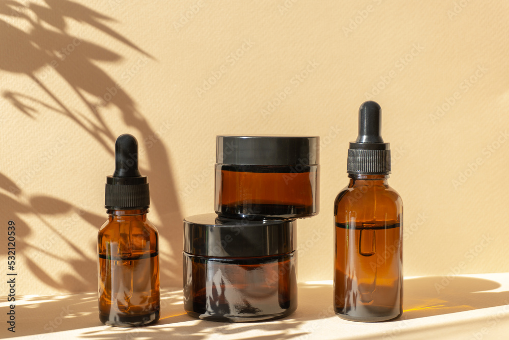 Mockup of amber-colored glass vials with a dropper lid, jars of cosmetic cream in sunlight on brown background with floral shadow. Concept of body and face care Mockup of amber-colored glass vials with a dropper lid, jars of cosmetic cream in sunlight on brown background with floral shadow. Concept of body and face care