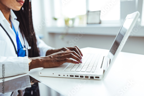 Close-up hands of unrecognizable female physician in medical uniform working typing on laptop keyboard sitting at desk on background of window in dark office room.