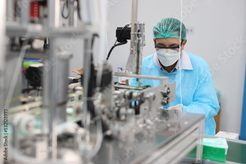 Worker in personal protective equipment or PPE inspecting quality of mask and medical face mask production line in factory, manufacturing industry and factory concept.