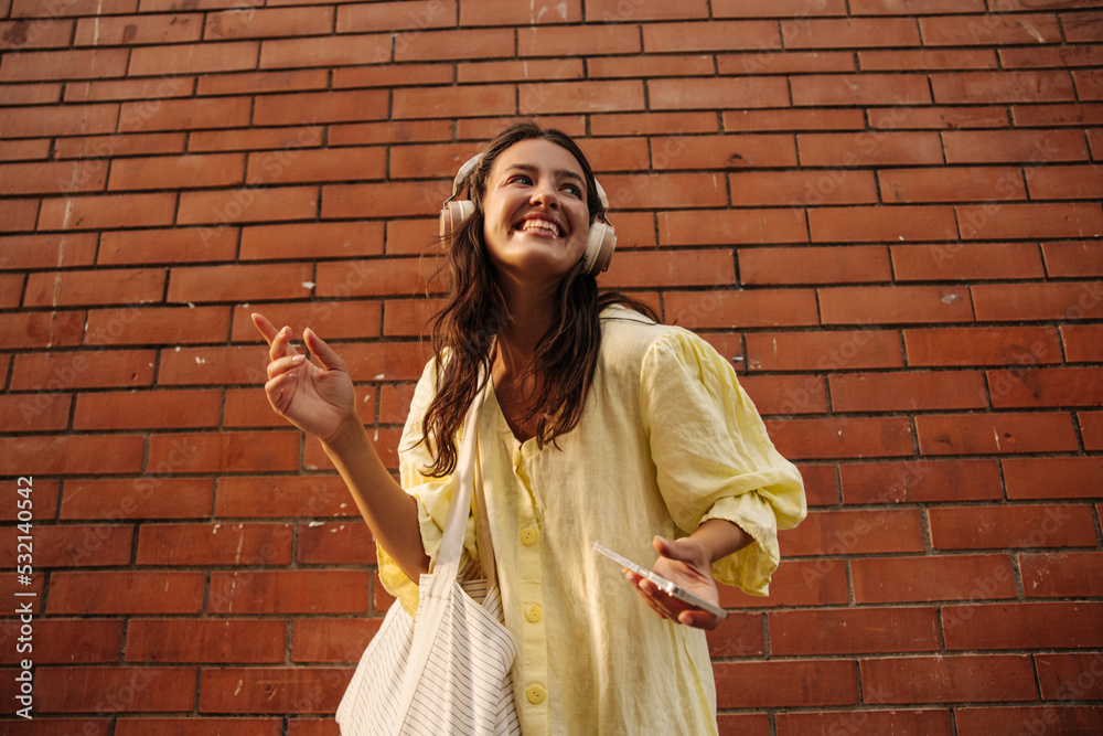 Positive young caucasian woman enjoys listening to music in her spare time outdoors. Brunette in dress wears headphones, phone and bag. Audio technology, lifestyle concept