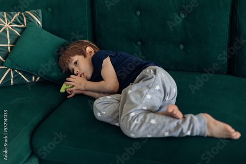 Portrait of upset, sad and depressed barefoot little boy lying on green sofa, sucking thumb finger. Psychological stress photo