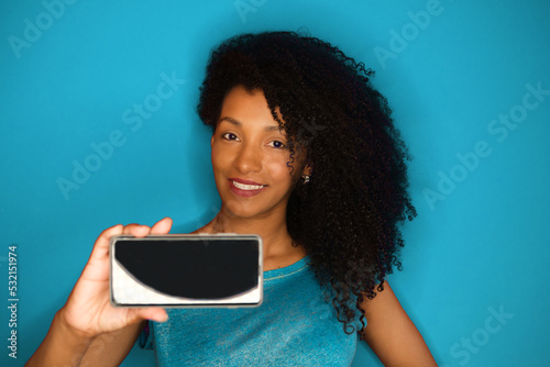 Charming beautiful black woman showing smartphone screen. Cheerful afro hairstyle model on blue background.