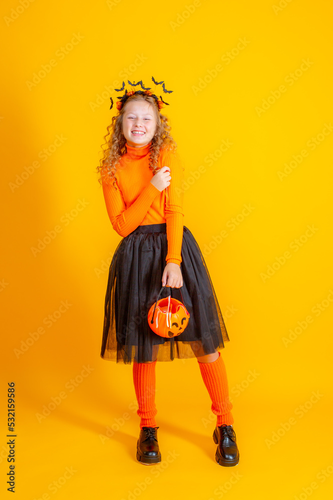 a teenage girl in a witch costume on a yellow background, holding a confent pumpkin eating marmalade worms halloween party