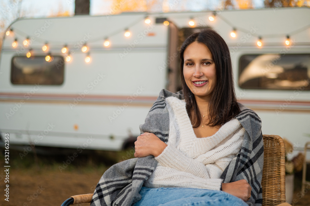 Caucasian woman sits in a wicker chair wrapped in a blanket in the yard near the trailer in autumn. Caucasian woman sits in a wicker chair wrapped in a blanket in the yard near the trailer in autumn.