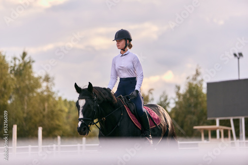 Young sportive girl, professional jockey or horsewoman in sports uniform and helmet riding black horse at sports arena. Horseback riding © master1305