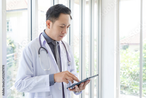 Portrait of asian senior doctor standing and reading in medical office while using tablet.