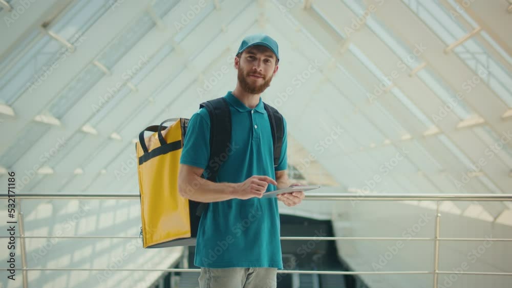 Food Delivery Person in Uniform and With a Thermal Backpack Walking in Modern Office Building and Using Tablet to Check Order. Courier On the Way to Deliver Order to a Client. Food Delivery Person in Uniform and With a Thermal Backpack Walking in Modern Office Building and Using Tablet to Check Order. Courier On the Way to Deliver Order to a Client.