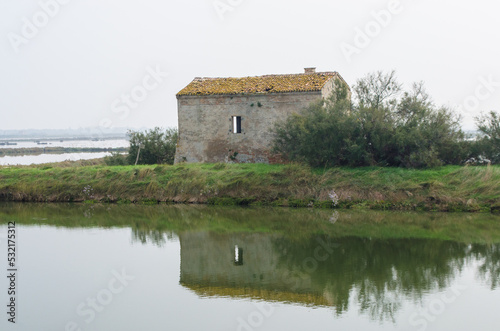 Un vecchio casone da pesca su una lingua di terra delle Valli di Comacchio si riflette in un canale in una giornata nuvolosa © Andrea Vismara