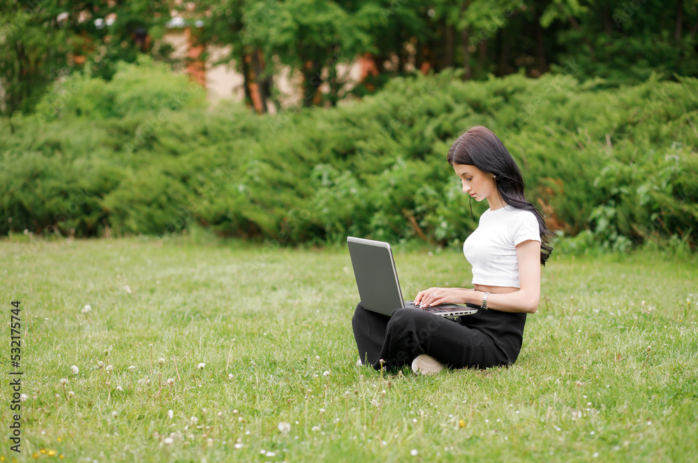 Portrait of a young woman sitting on green grass in a park with legs crossed on a summer day while using a laptop Portrait of a young woman sitting on green grass in a park with legs crossed on a summer day while using a laptop