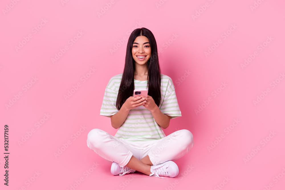 Full length photo of positive adorable indonesian person sit floor use telephone isolated on pink color background Full length photo of positive adorable indonesian person sit floor use telephone isolated on pink color background