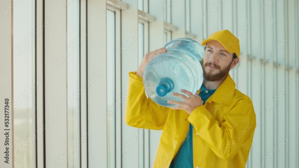 Handsome Delivery Man in Yellow Uniform Brings on Shoulder a Big Bottle of Water at Modern Office Building.Courier on the Way to Deliver Order to Client. Deliveryman Worker Deliver Online Order Client Handsome Delivery Man in Yellow Uniform Brings on Shoulder a Big Bottle of Water at Modern Office Building.Courier on the Way to Deliver Order to Client. Deliveryman Worker Deliver Online Order Client