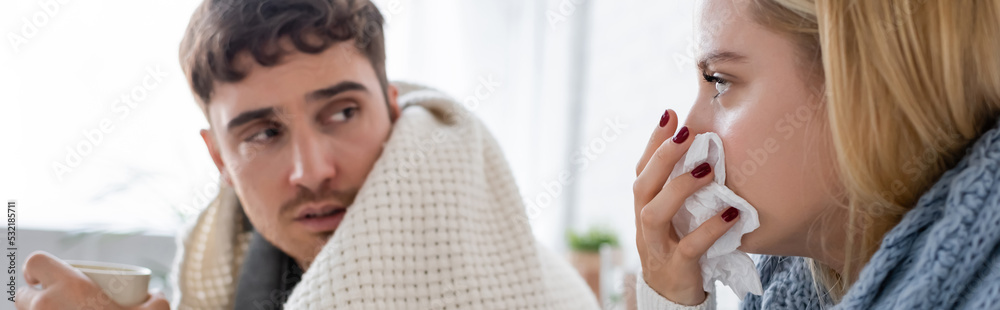 Obraz premium sick blonde woman in scarf sneezing in tissue near diseased boyfriend with cup of tea, banner.