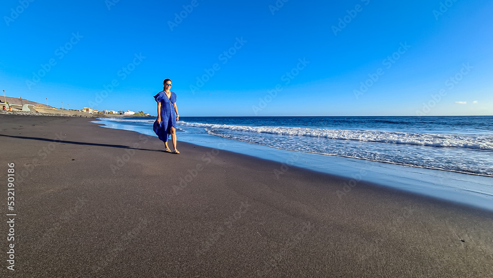 Happy luxury woman in dress walking on the beach Playa Valle Gran Rey,  Promenade La Calera in Valle Gran Rey on La Gomera, Canary Islands, Spain, Europe. Dark volcanic sand beach. Flower in hair