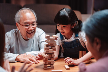 Asian happy family of little daughter play toy wooden block in home. Grandparents and young woman loving couple parent having fun play stack tower wood block game at night in living room.