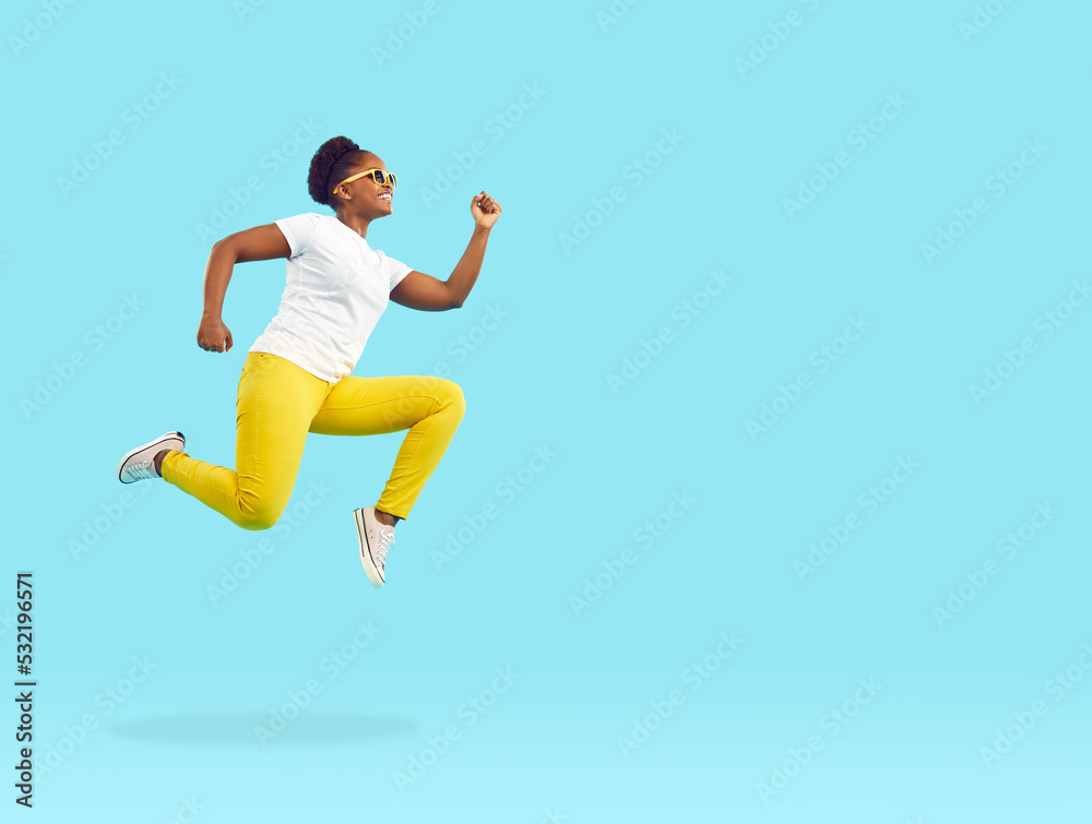 Side view happy energetic young African American woman in white T shirt, yellow trousers and sunglasses running, flying or jumping high in air isolated on bright blue blank empty copy space background Side view happy energetic young African American woman in white T shirt, yellow trousers and sunglasses running, flying or jumping high in air isolated on bright blue blank empty copy space background