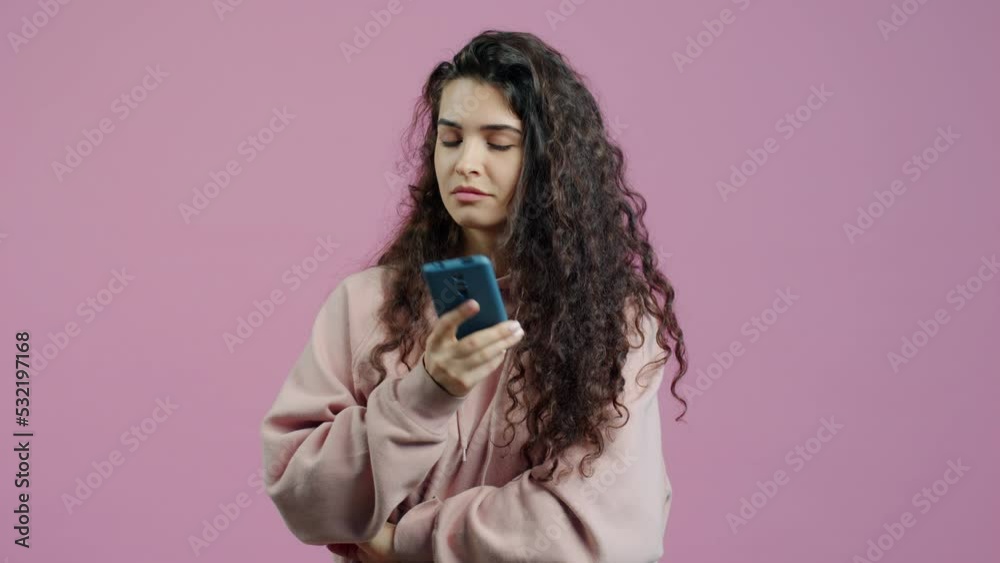 Smiling young lady is using smartphone texting and looking at camera standing on pink background alone. Communication and lifestyle concept. Smiling young lady is using smartphone texting and looking at camera standing on pink background alone. Communication and lifestyle concept.