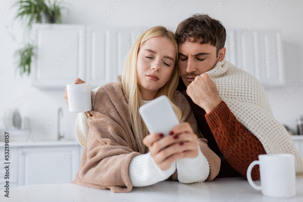 blonde young woman using mobile phone near boyfriend and cup on table. blonde young woman using mobile phone near boyfriend and cup on table.