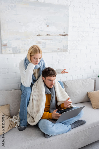 displeased blonde woman in sweater and scarf sitting near freelancer and gesturing while looking at laptop.