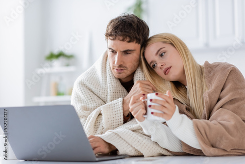 young blonde woman covered in blanket leaning on shoulder of boyfriend using laptop and working from home.