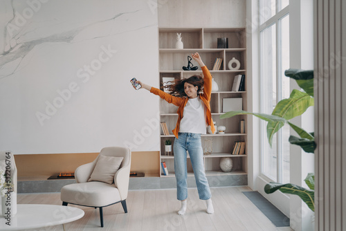 Energetic woman dancing in living room by music from smartphone in modern apartment home interior photo