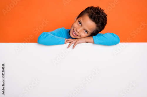 Photo portrait of adorable small pupil boy hide behind blank ad print space dressed stylish blue look isolated on orange color background