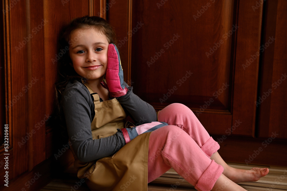 Portrait of an adorable Caucasian child girl, little baker confectioner in a beige chef apron and kitchen mittens, sitting on the floor and leaning against wooden kitchen cupboard, smiling to camera