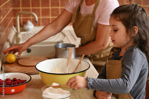 Beautiful little girl, adorable lovely daughter in chef's apron, adding scoop of sugar into a bowl with dry ingredients, preparing dough with her mother in home kitchen. Culinary and baking concept