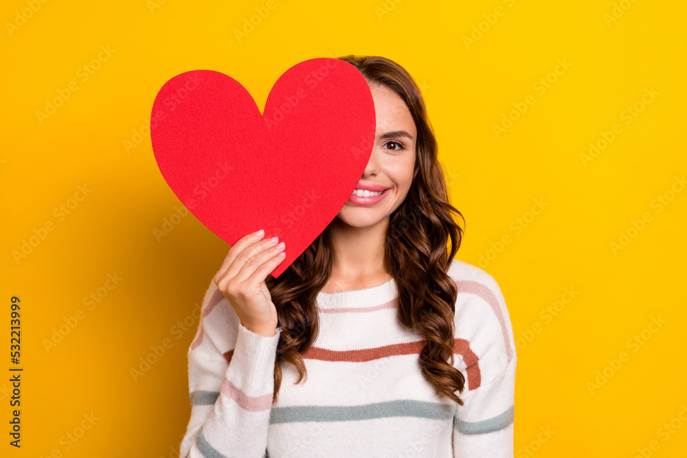 Portrait of cheerful cute girl hand hold heart symbol card cover one eye isolated on yellow color background Portrait of cheerful cute girl hand hold heart symbol card cover one eye isolated on yellow color background