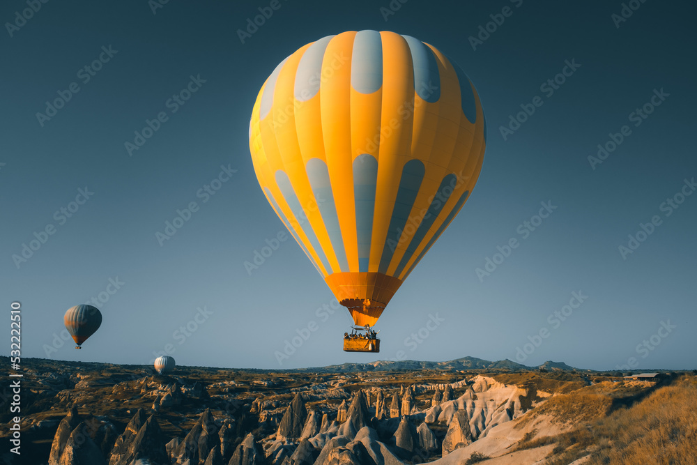 Obraz premium Yellow bright hot air balloon with tourists rise up for sunrise watching. Wide blue sky landscape of Goreme valley in Cappadocia. Tourism, travel, holidays, recreation background, concept postcard