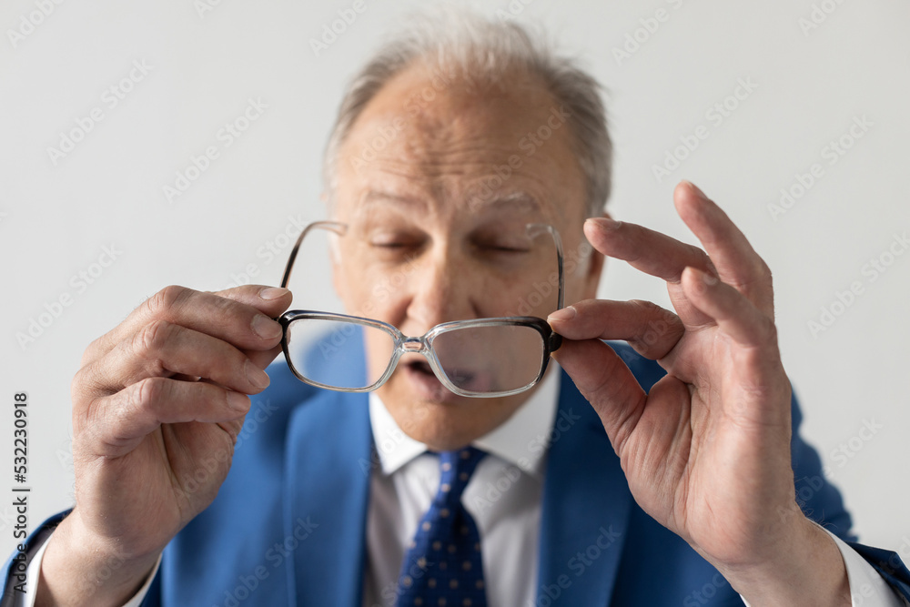 Close-up of short-sighted mature businessman looking at glasses. Portrait of senior manager wearing formalwear holding spectacles against white background. Optometry concept