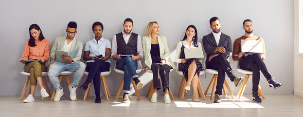 Diverse mixed race people reading or using different devices not to get bored while sitting on chairs in office corridor and waiting in line for their appointment, job interview, or business meeting